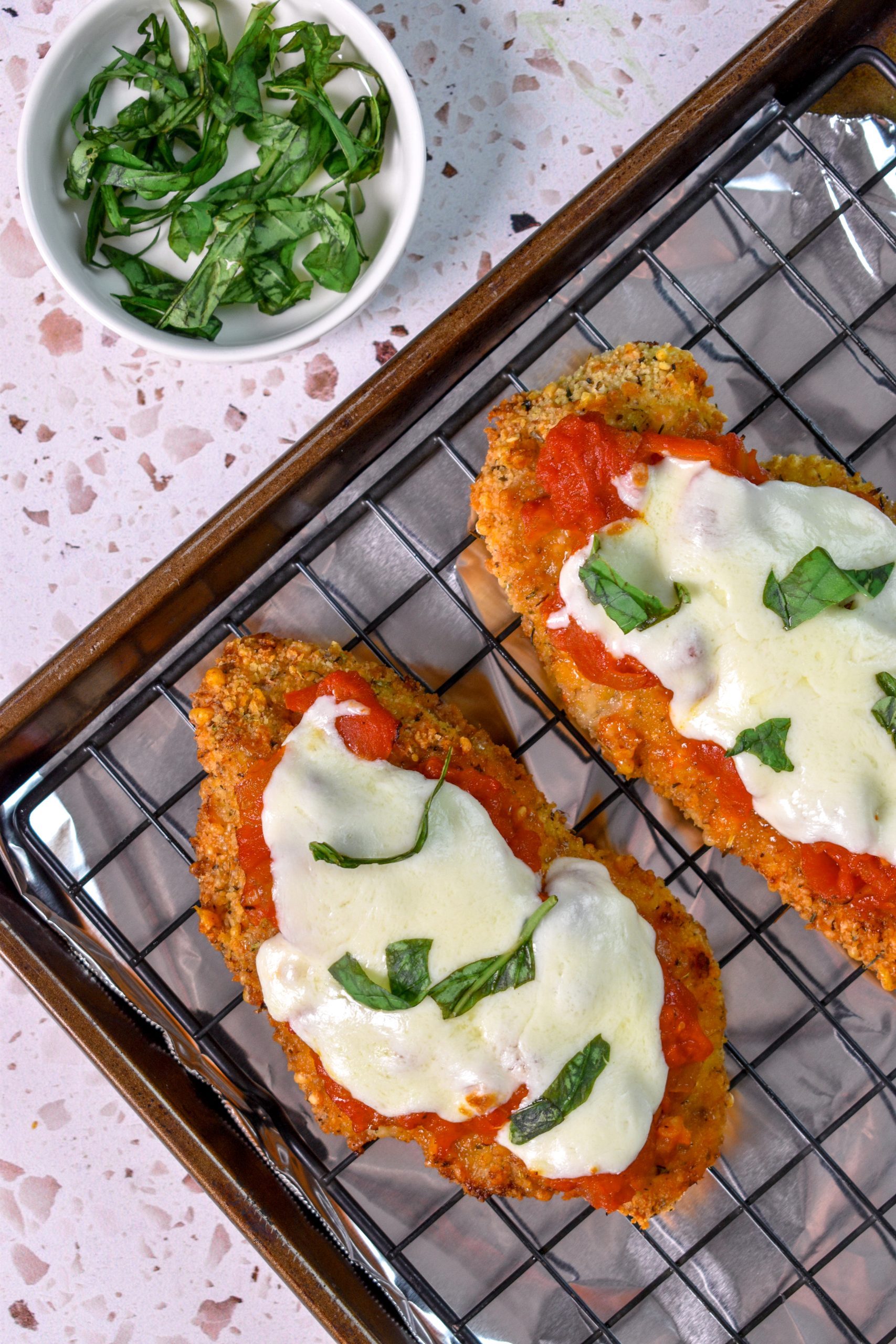 This is an image of air fried chicken parmesan sitting on a cooling rack that's on a baking sheet there is a bowl of basil on the side