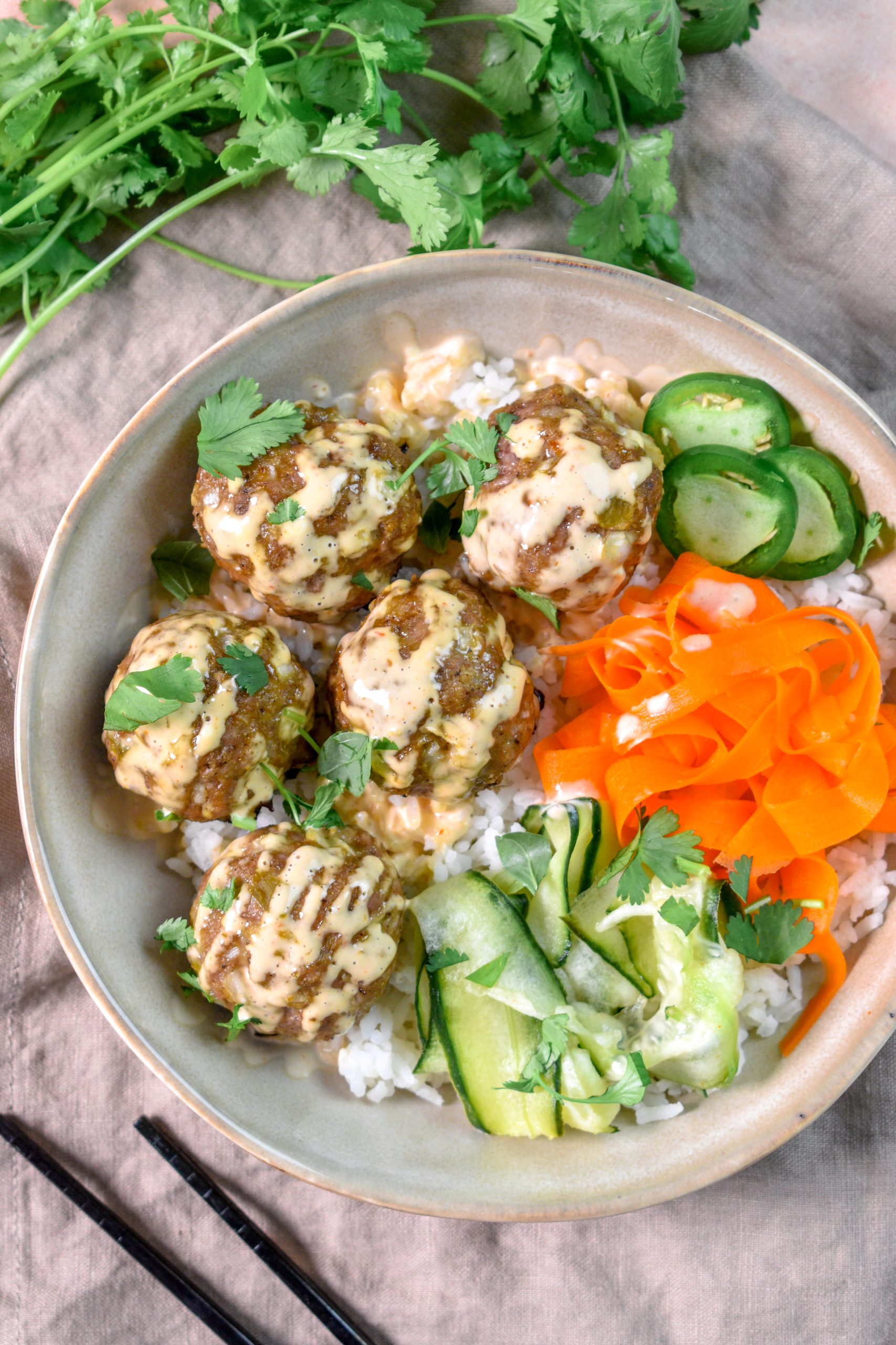 This is an image of a bowl of banh mi meatballs sitting on a bed of rice with a side of pickled cucumber and carrots and topped with fresh jalapenos and cilantro