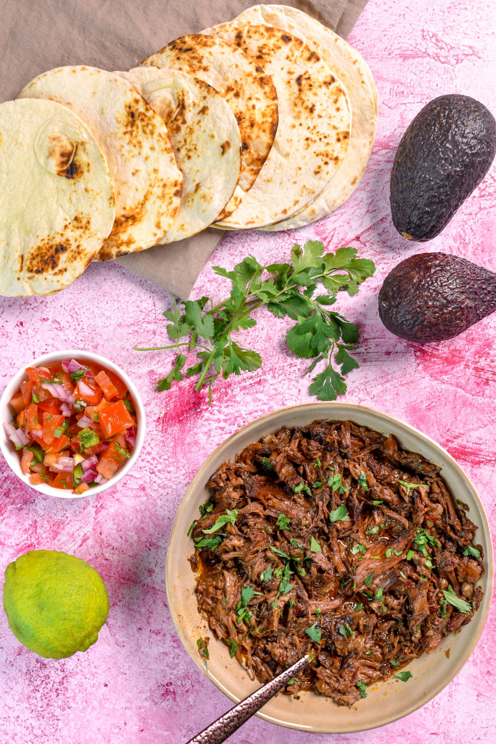 This is an image of beef barbacoa in a brown bowl. Around it is a lime, pico de gallo, two avocados, cilantro, and tortillas