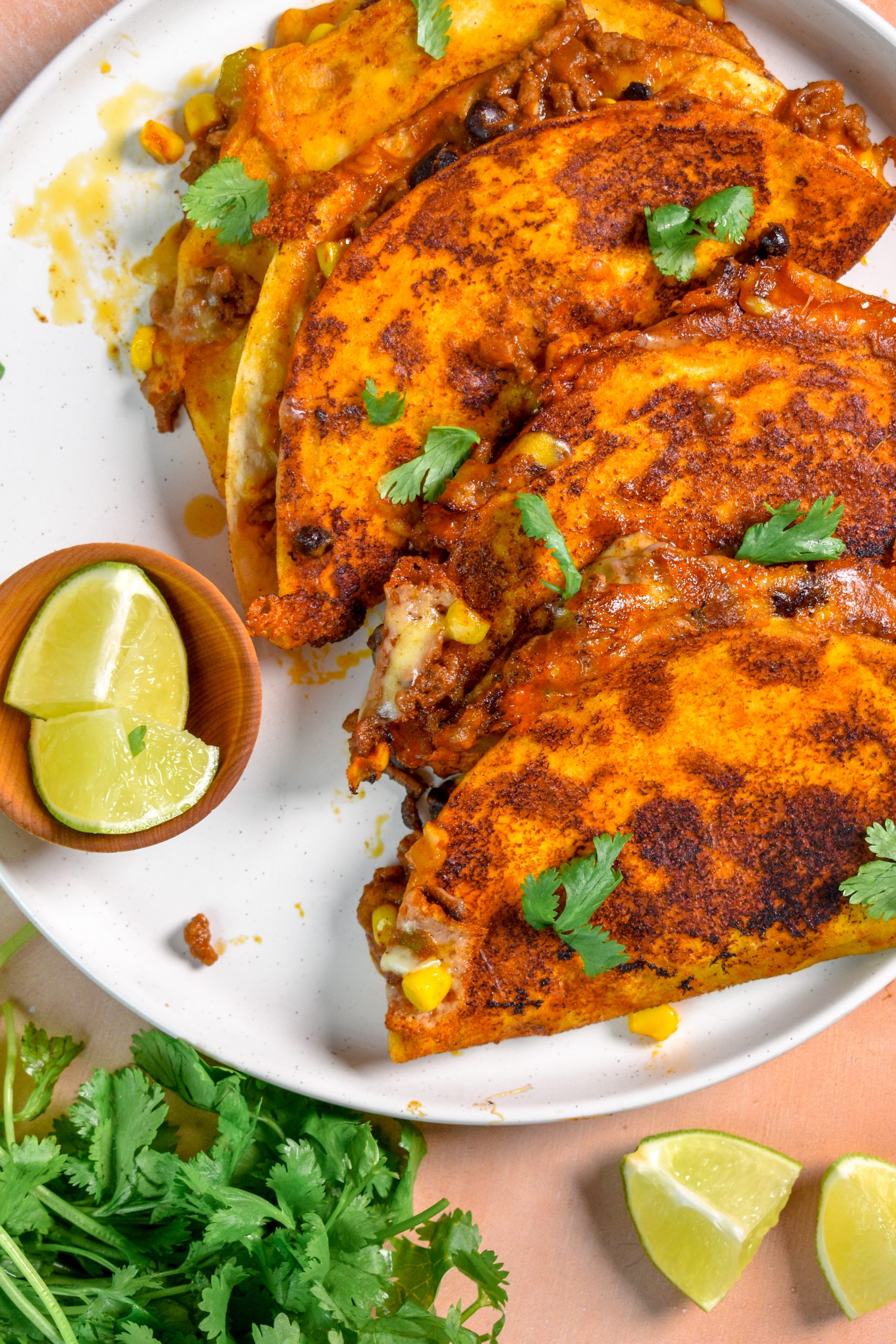 This is an image of enchilada tacos sitting on a white plate with lime wedges and cilantro.