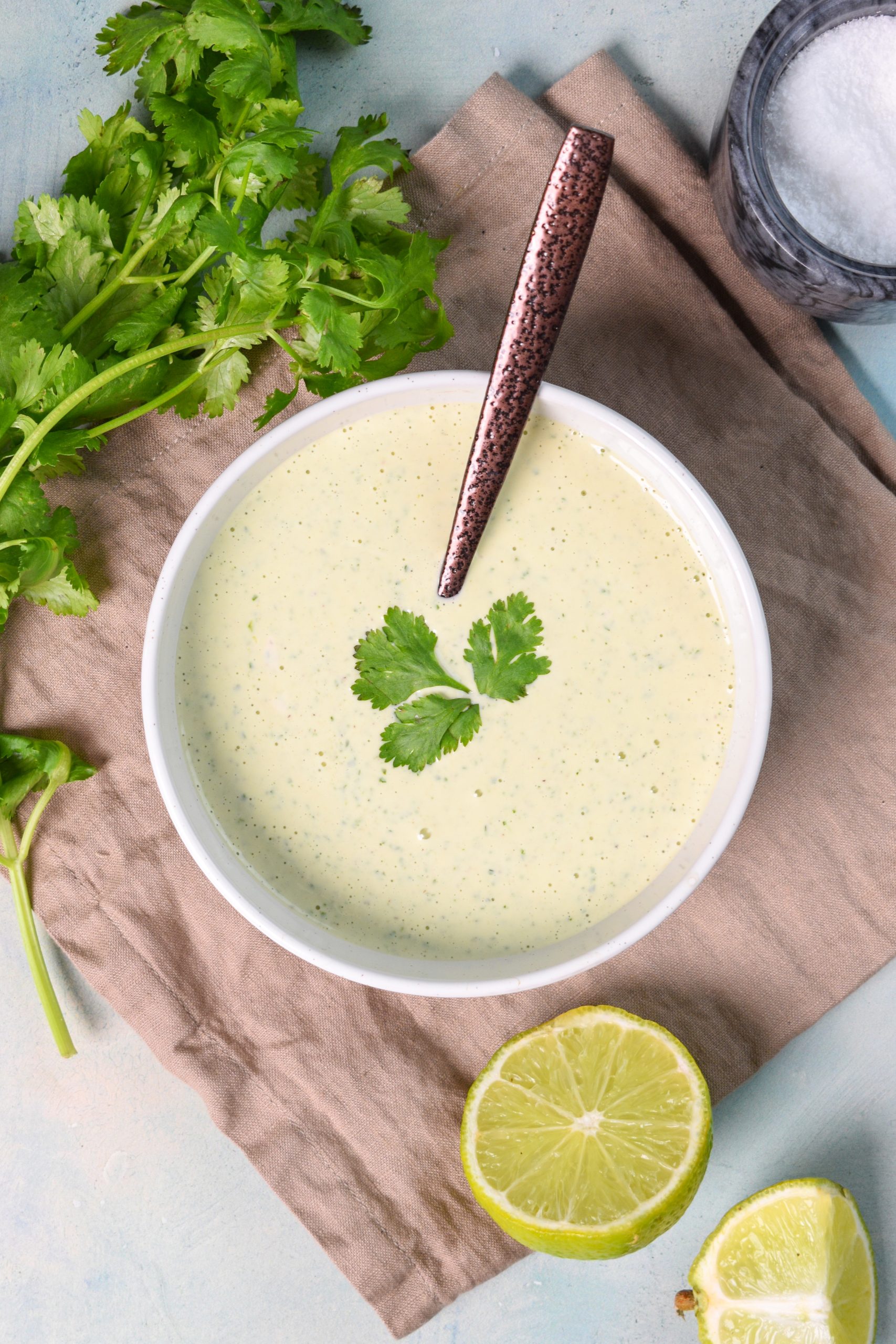 This is an image of cilantro lime dressing in a white bowl with a spoon resting in it. The bowl is sitting on top of a tan towel and there's cilantro, limes, and salt around the bowl.