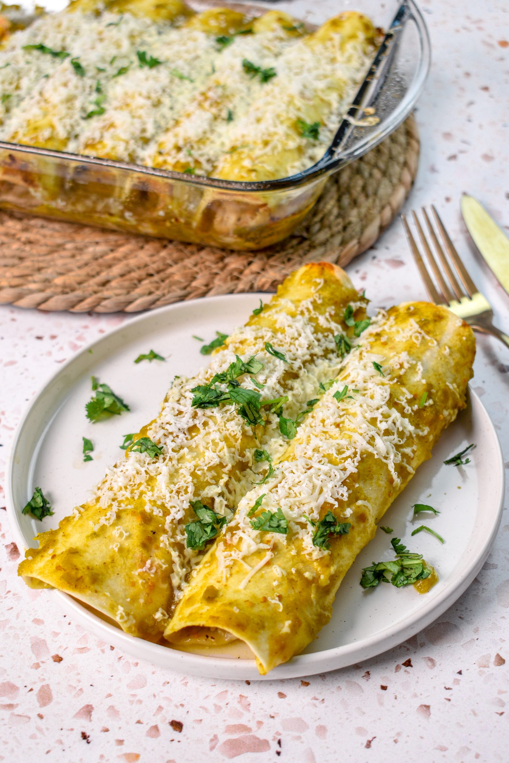 This is an image of elote chicken enchiladas sitting on a white plate with more enchiladas in a casserole dish in the background.