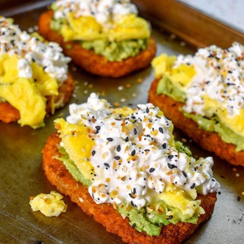 This is an image of 4 hash browns sitting on a pan topped with avocado, eggs, cottage cheese, and bagel seasoning