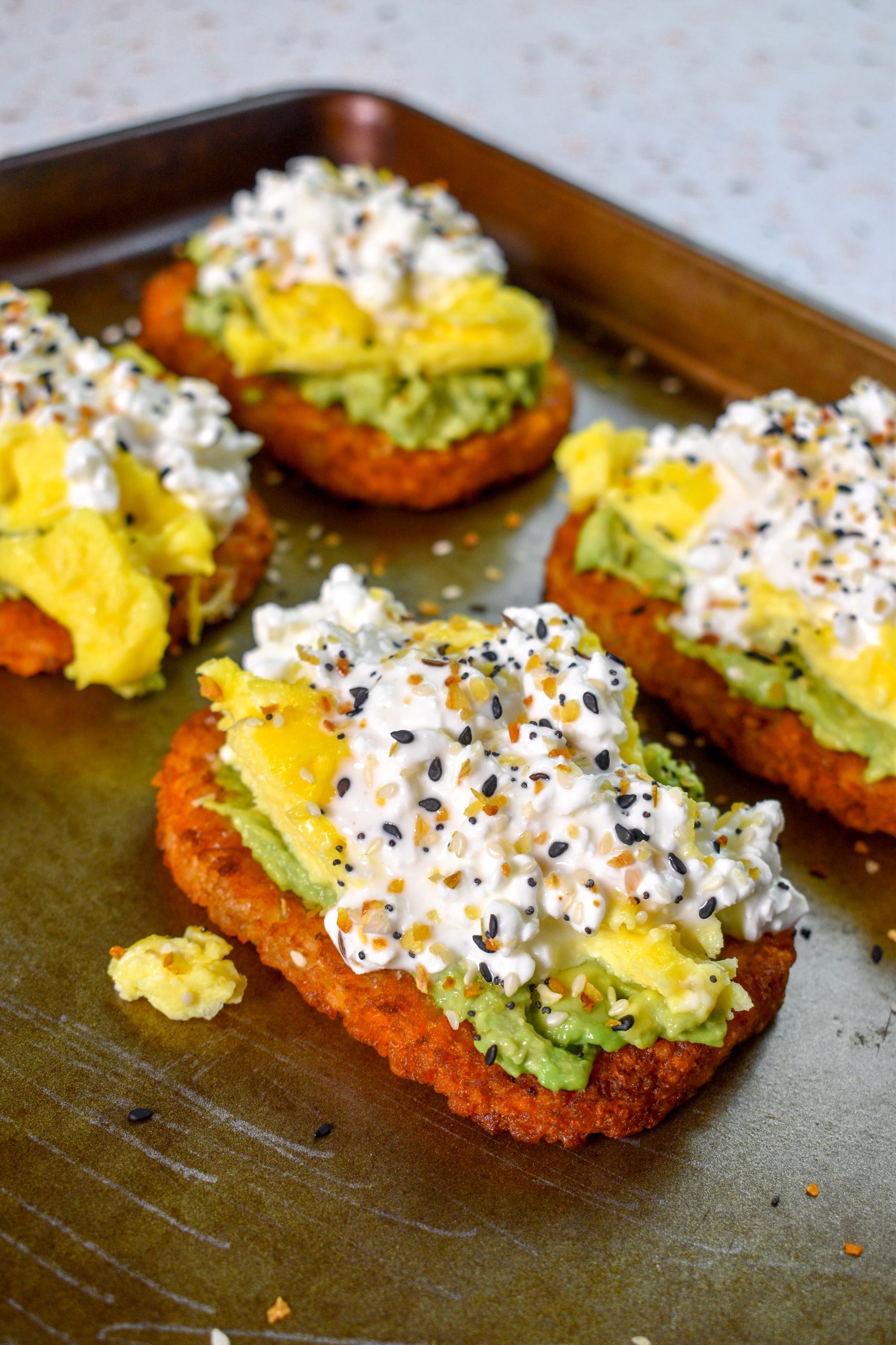 This is an image of 4 hash browns sitting on a pan topped with avocado, eggs, cottage cheese, and bagel seasoning