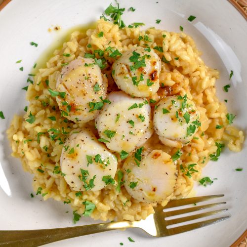 This is an image of seared scallops over lemon parmesan risotto sitting in a white bowl with a fork resting in the bowl.