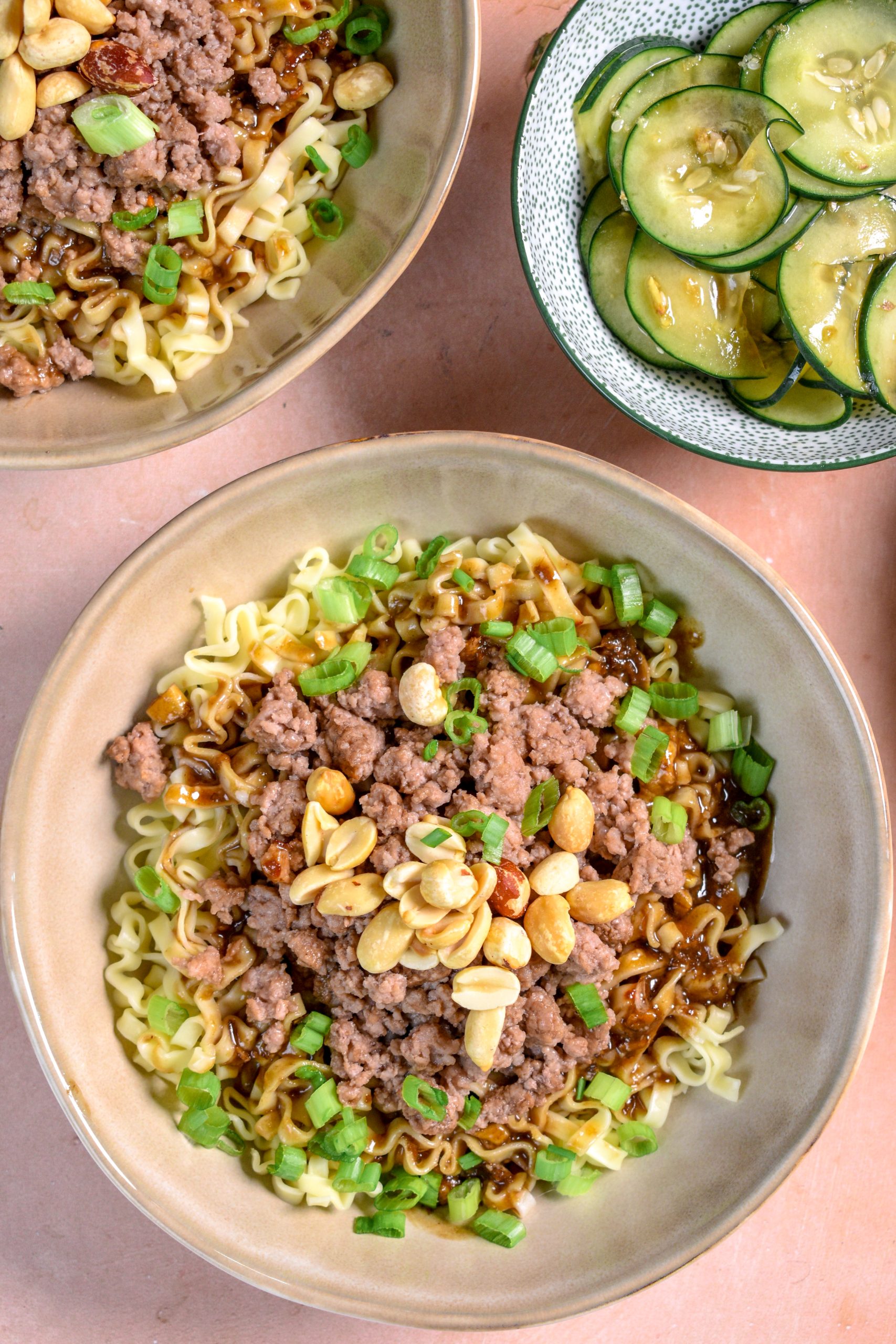 This is an image of dan dan noodles sitting in a bowl topped with ground pork, peanuts and scallions. There is also a bowl of cucumber salad on the side.