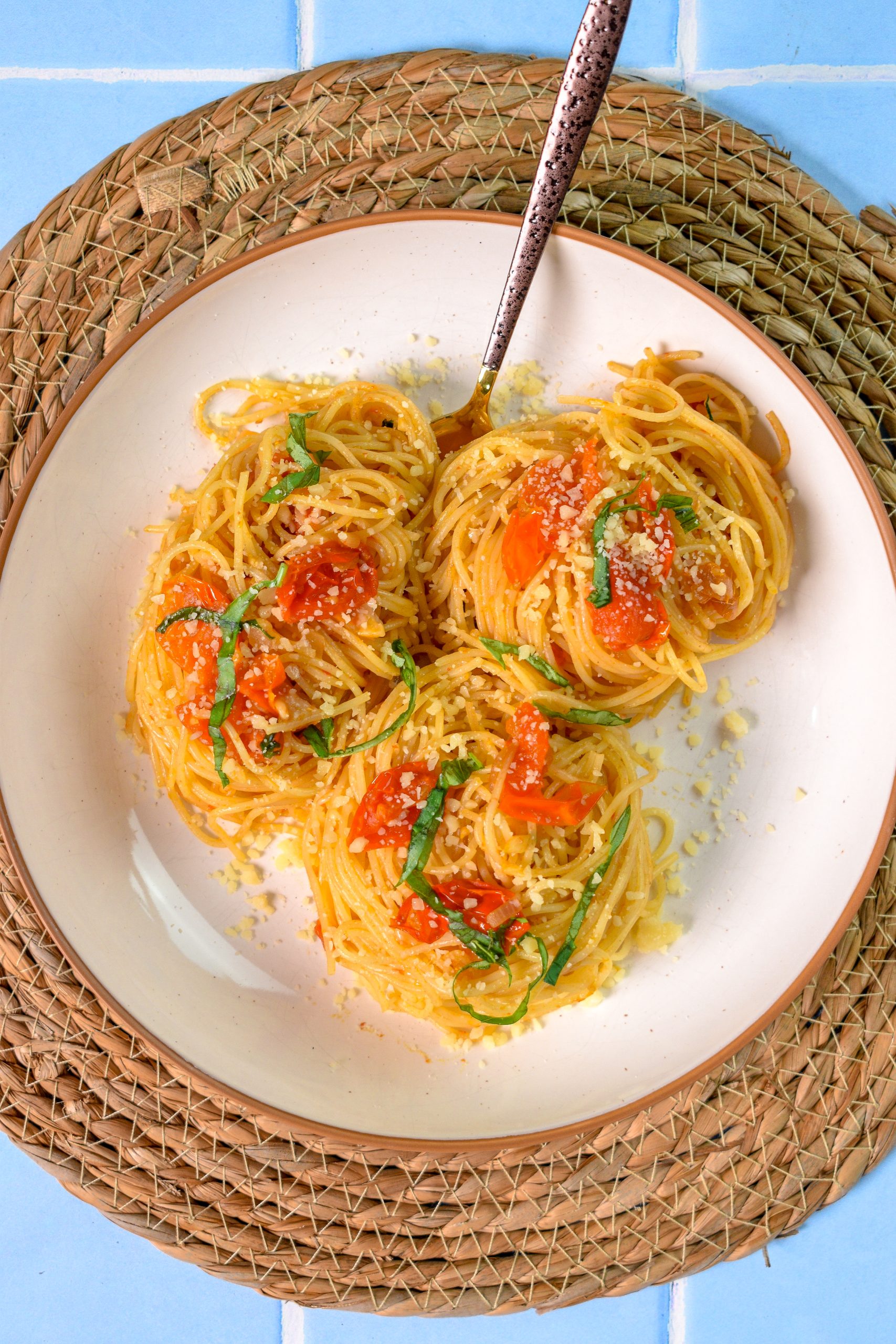 This is an image of anchovy tomato pasta sitting in a white bowl with a spoon resting in it. The bowl is sitting on top of a wicker charger.