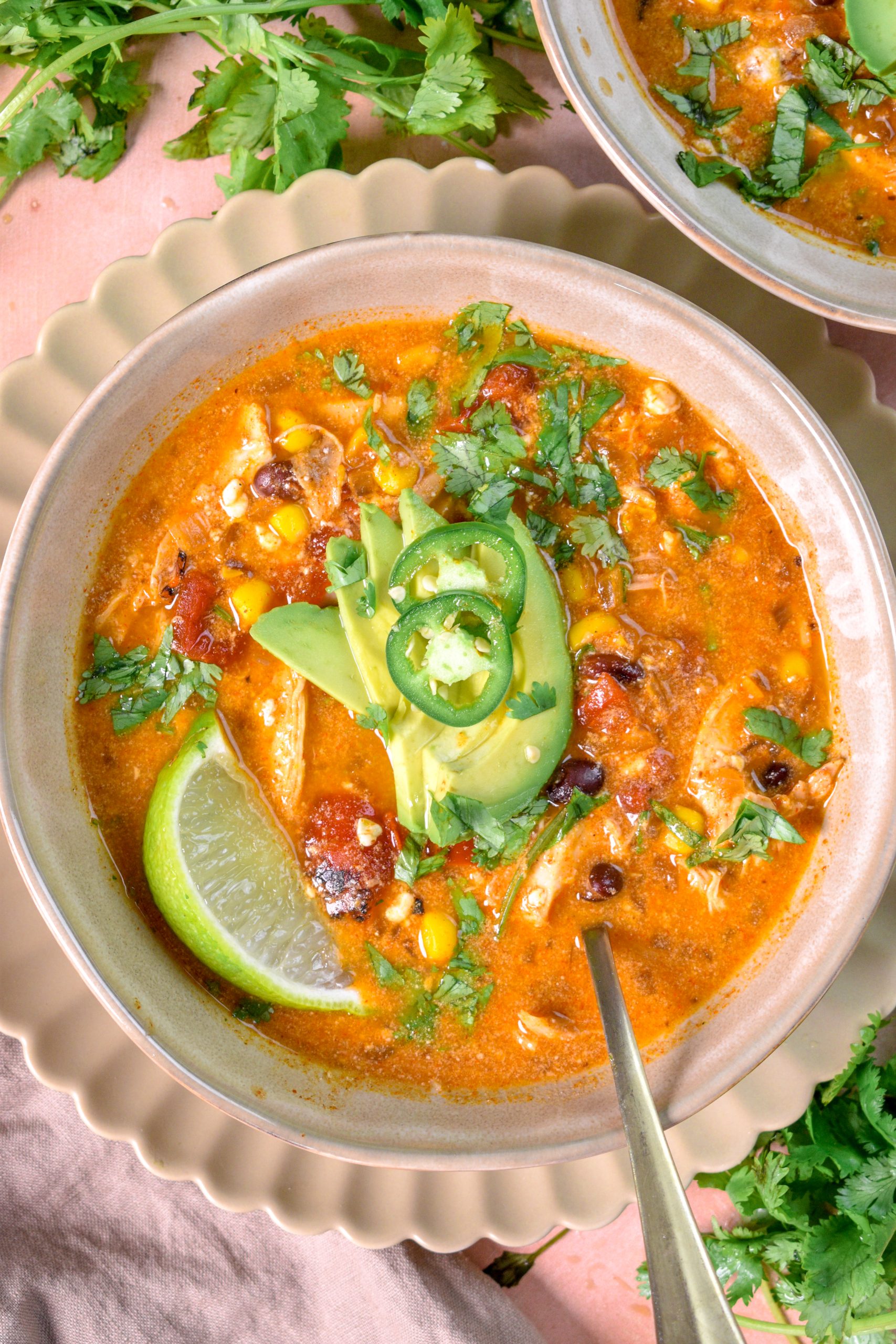 This is an image of slow cooker high protein chicken enchilada soup sitting in a bowl topped with avocado and jalapeno. There's a spoon resting in the bowl and cilantro around it as well.