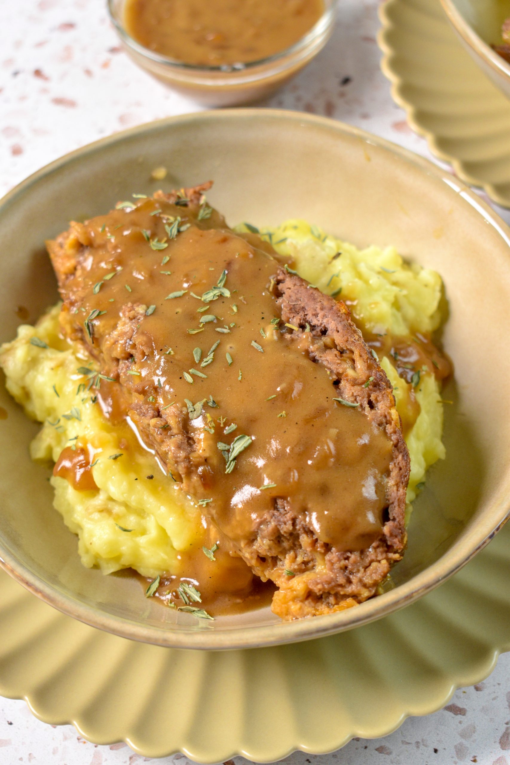 This is an image of french onion meatloaf sitting on a bed of mashed potatoes that's in a bowl. The meatloaf is topped with gravy and some thyme. In the background is some more gravy in a bowl.