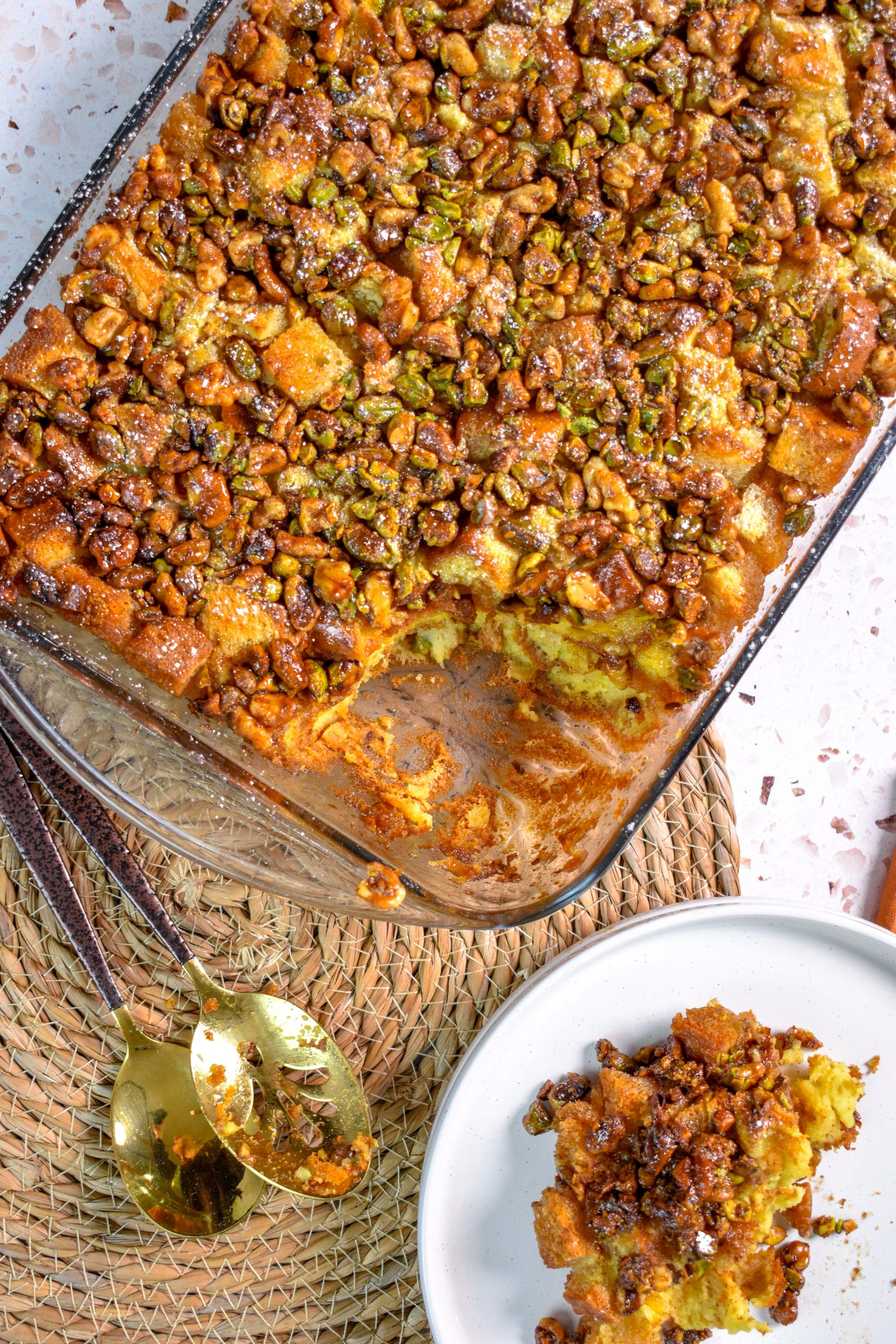 This is an image of baklava french toast bake in a casserole dish with a portion of it sitting on the side on a white plate. There are also 2 spoons resting to the side.