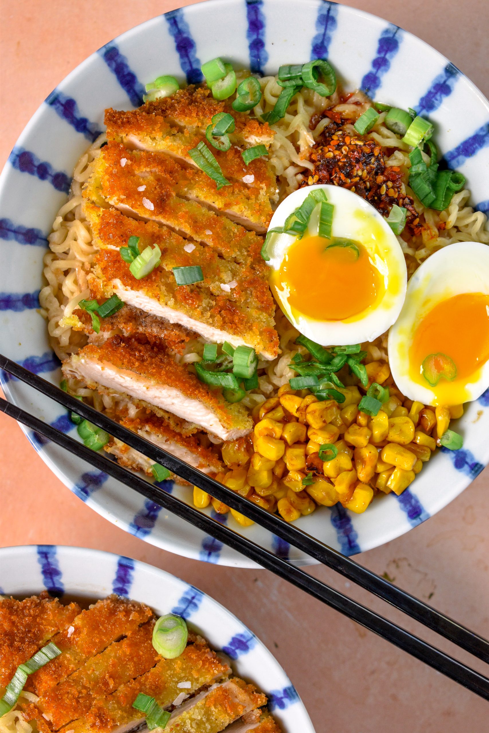 This is an image of chicken katsu ramen in a bowl topped with corn, chili oil, soft boiled eggs, and scallions