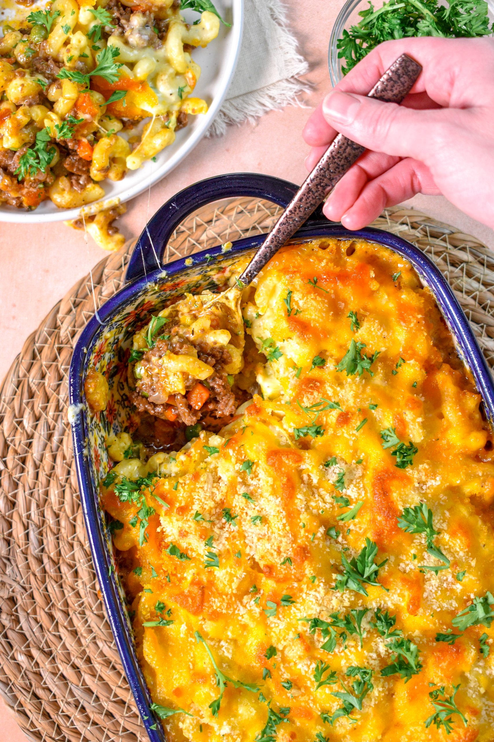 This is an image of Mac n Cheese Shepherds Pie. There is some sitting on a white plate and some is being spooned out of a casserole dish. There is also a bowl of parsley in the top right.