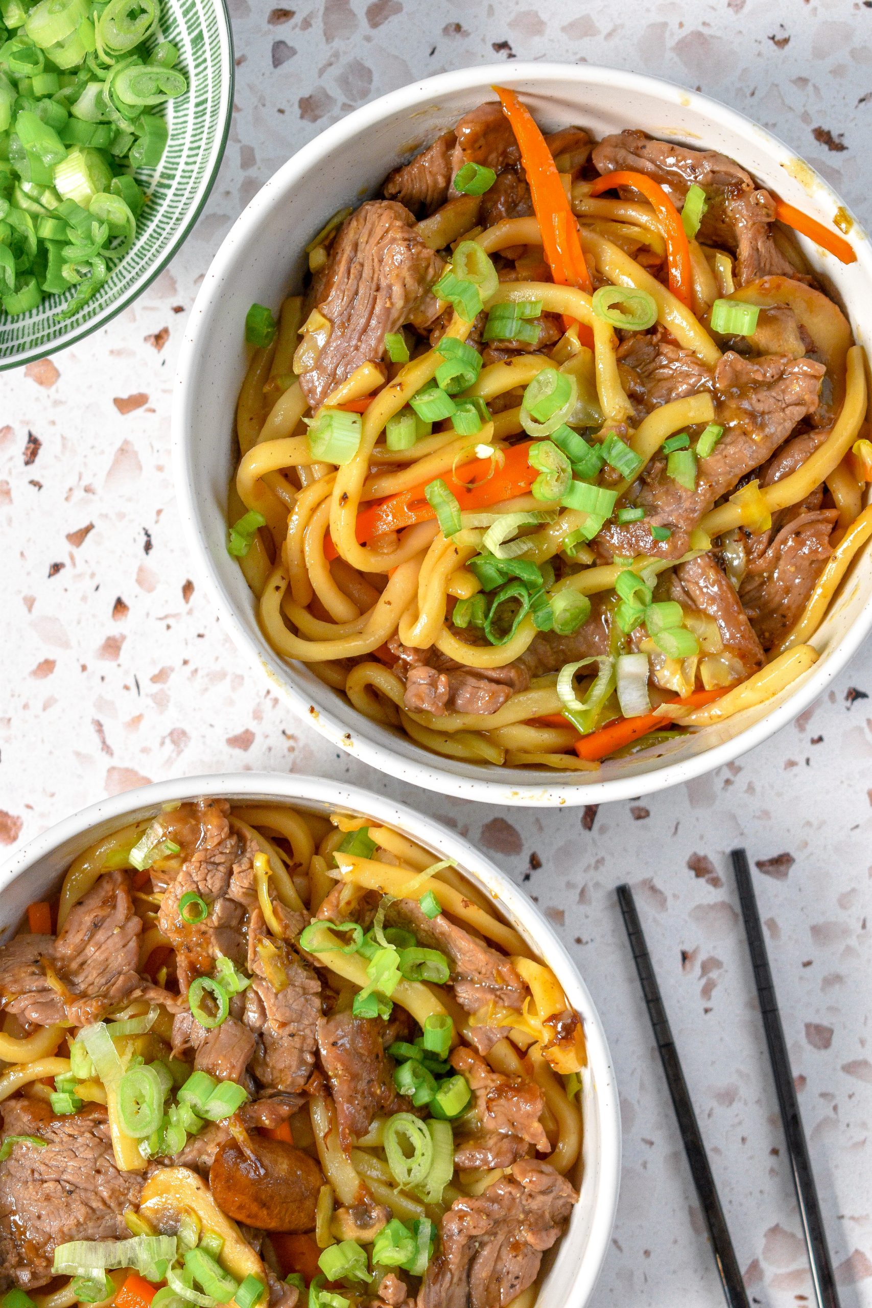 Two bowls of beef udon noodles loaded with vegetables and beef and garnished with green onions. There is also a bowl of green onions in the top left and in the bottom right are some chopsticks.