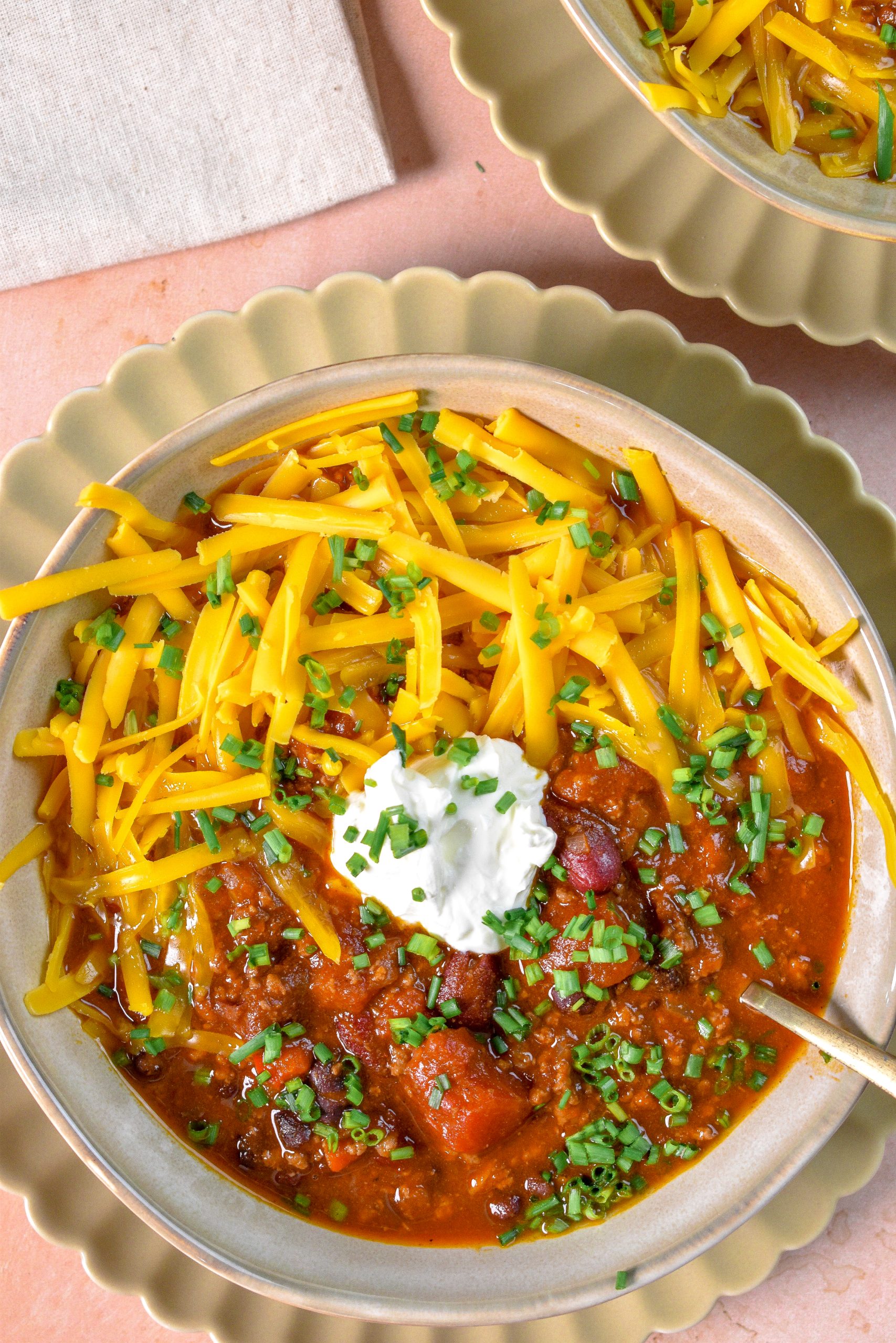 This is an image of Irish Chili in a tan bowl topped with shredded cheddar cheese, sour cream, and chives.