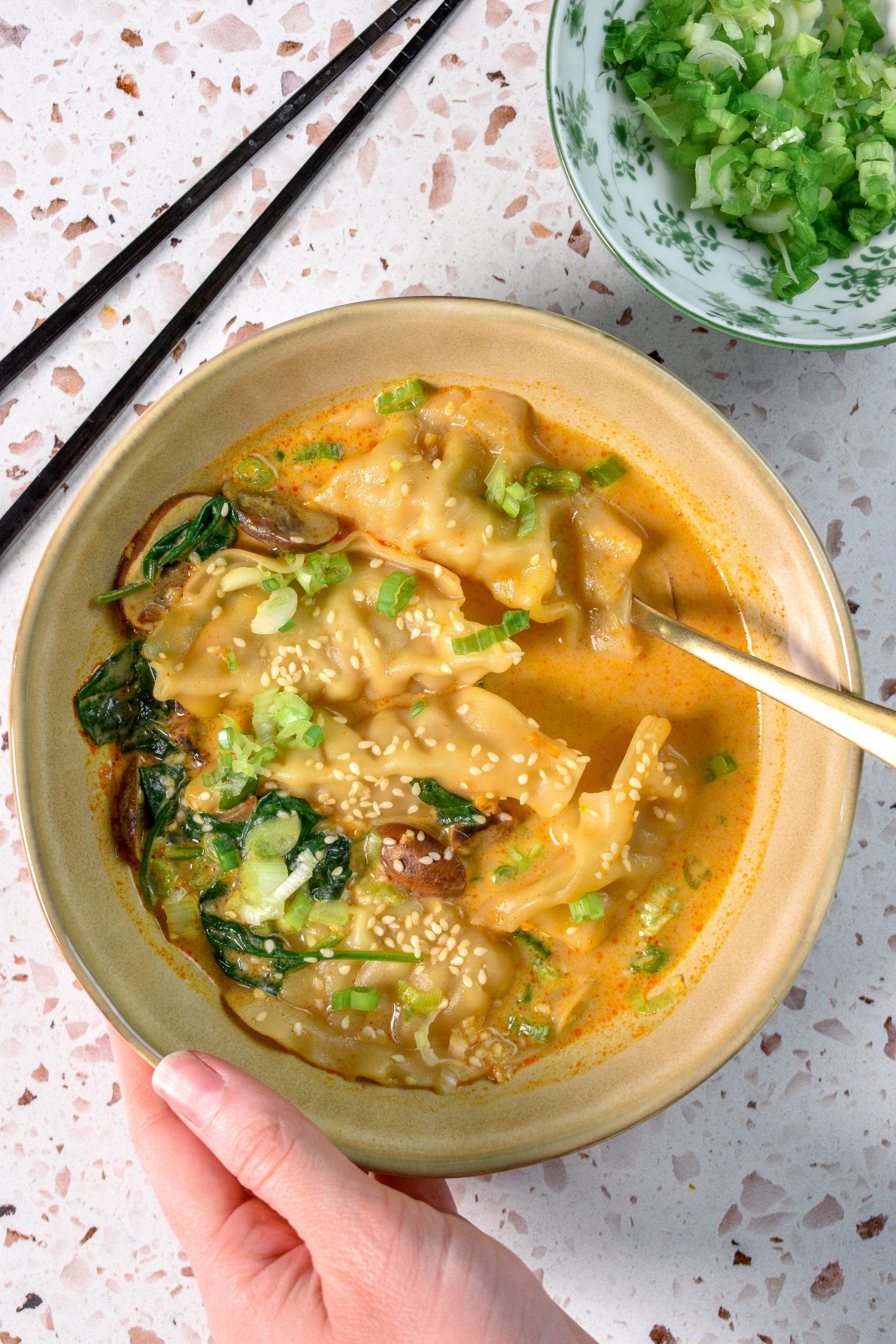 This is an image of Red Curry Potsticker Soup. There is a spoon resting in the dish and a hand holding the dish on the bottom. In the top right is a bowl of scallions and in the top left are some chopsticks.