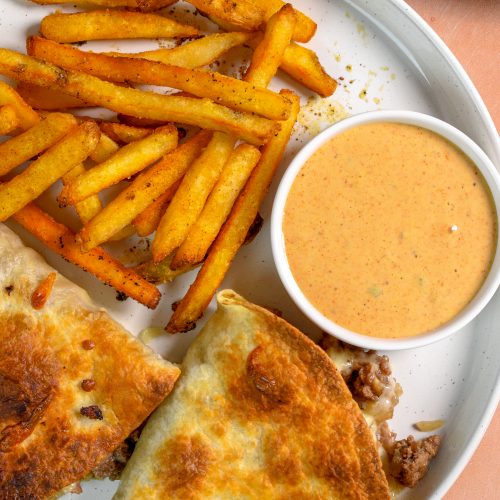 This is an image of tortilla cheeseburger pockets sitting on a white plate with fries and sauce on the side.
