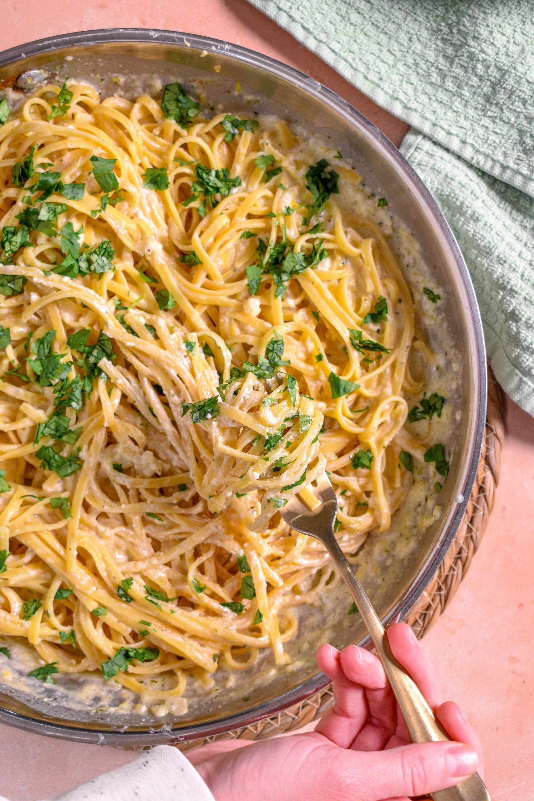 This is an image of creamy lemon boursin pasta in a stainless steel pan. There is a hand holding a fork with pasta on it and a green dish towel to the top right.